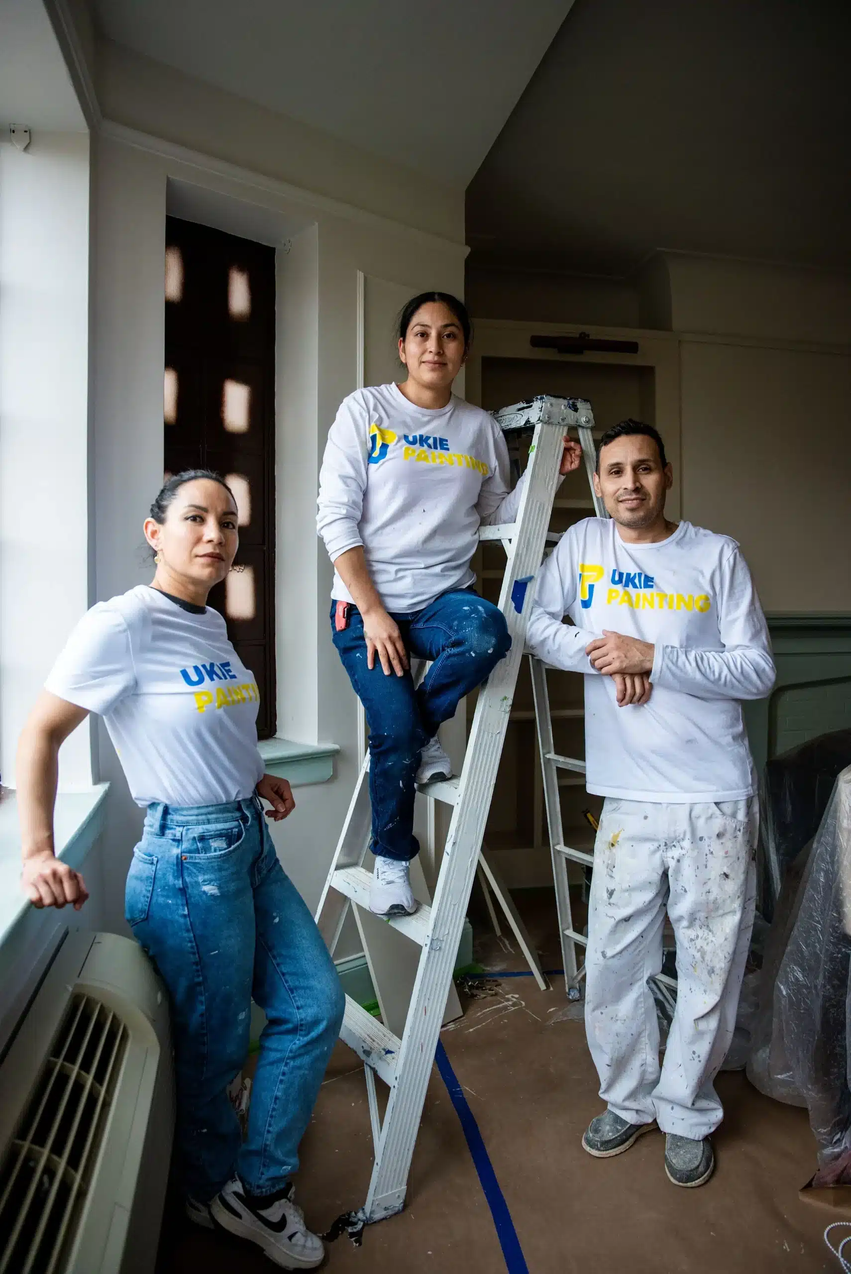 Our Skilled Painting Crew Ready for Your Interior Renovation Three professional Ukie Painting crew members, including one on a ladder, pose in a room prepared for painting.