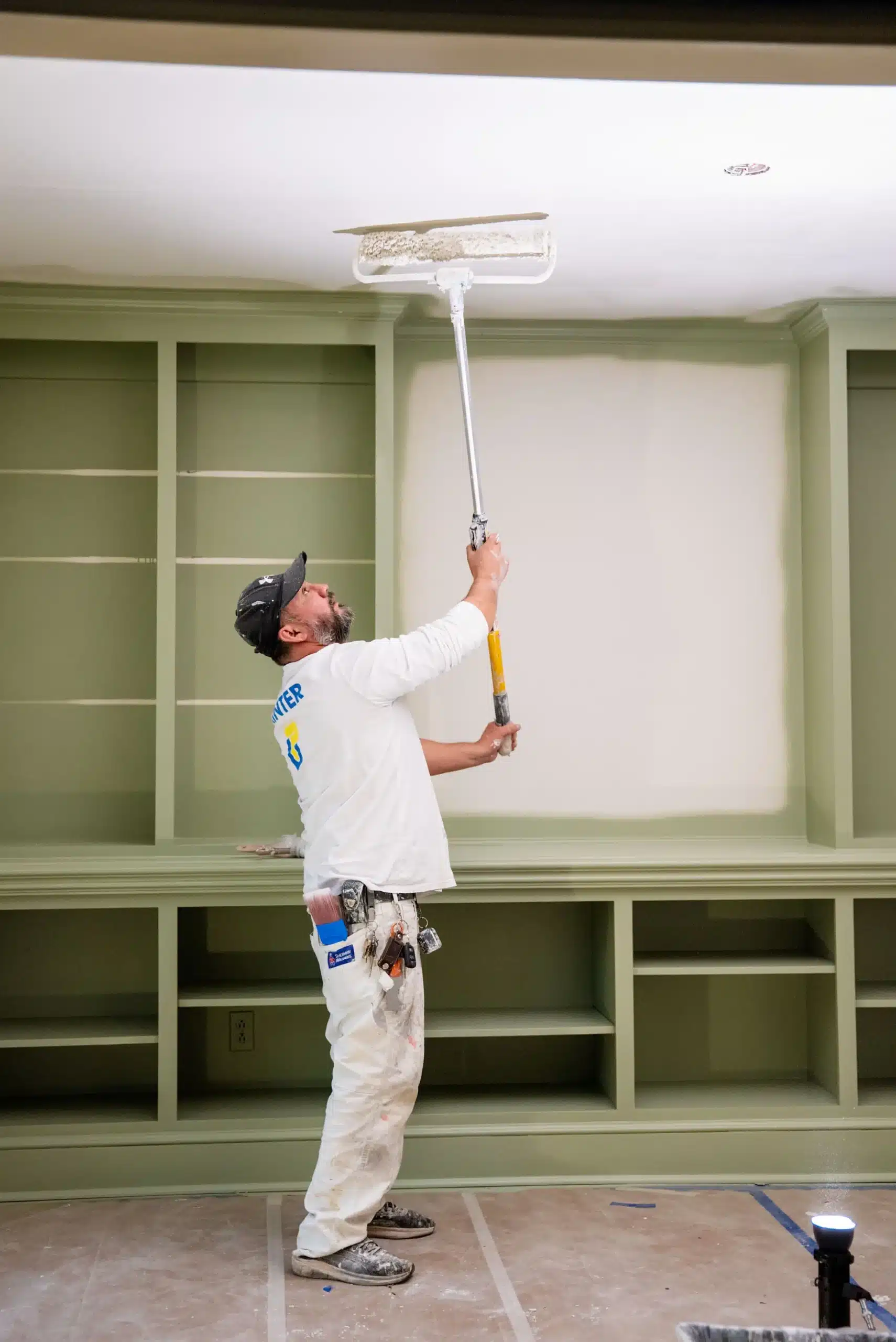 Professional Interior Ceiling Painting Services A painter using an extension roller to expertly paint the ceiling of a room with green built in shelves.