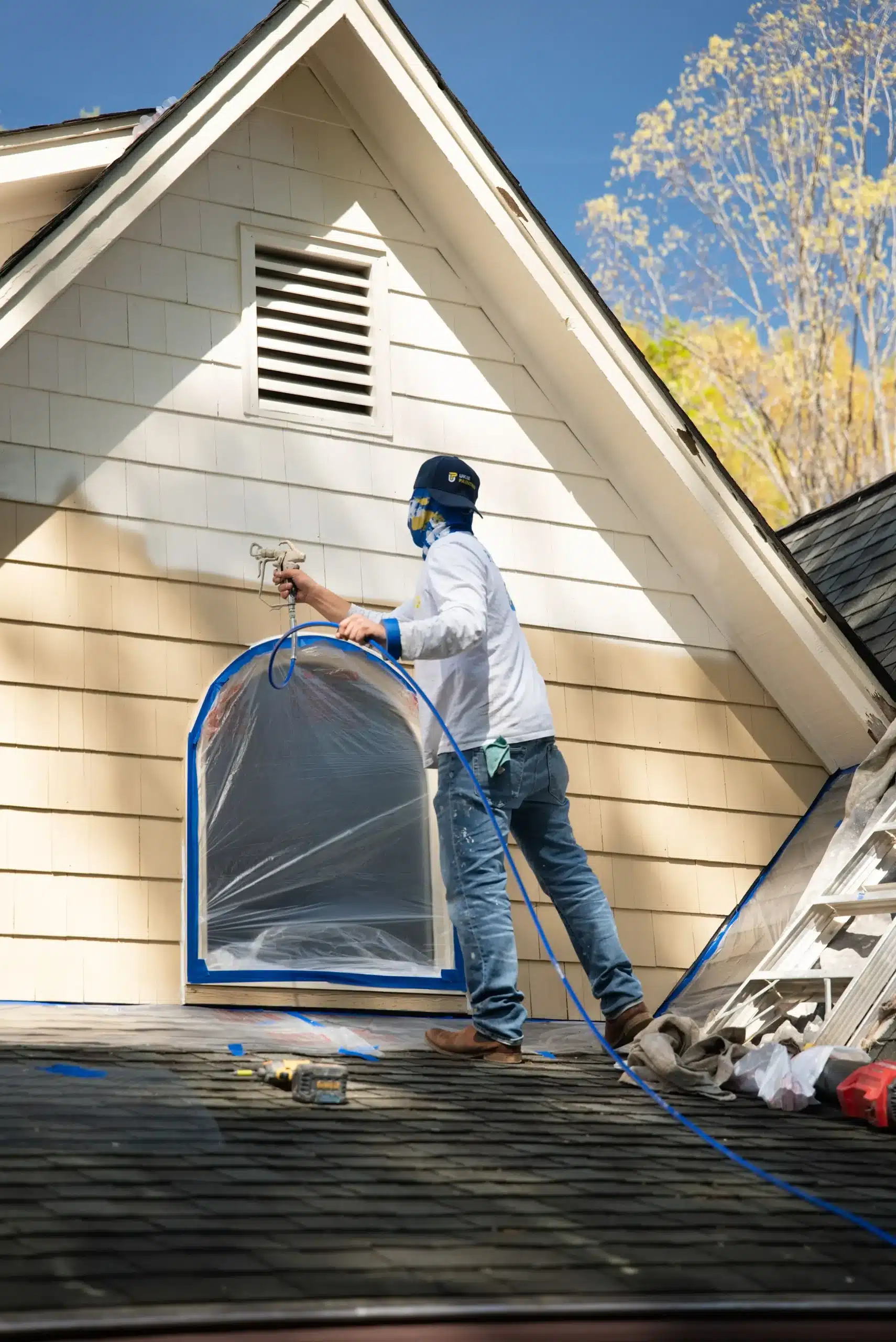Exterior House Painting with Professional Spray Application A painter on a roof spray painting the exterior siding of a house, with a protected arched window.