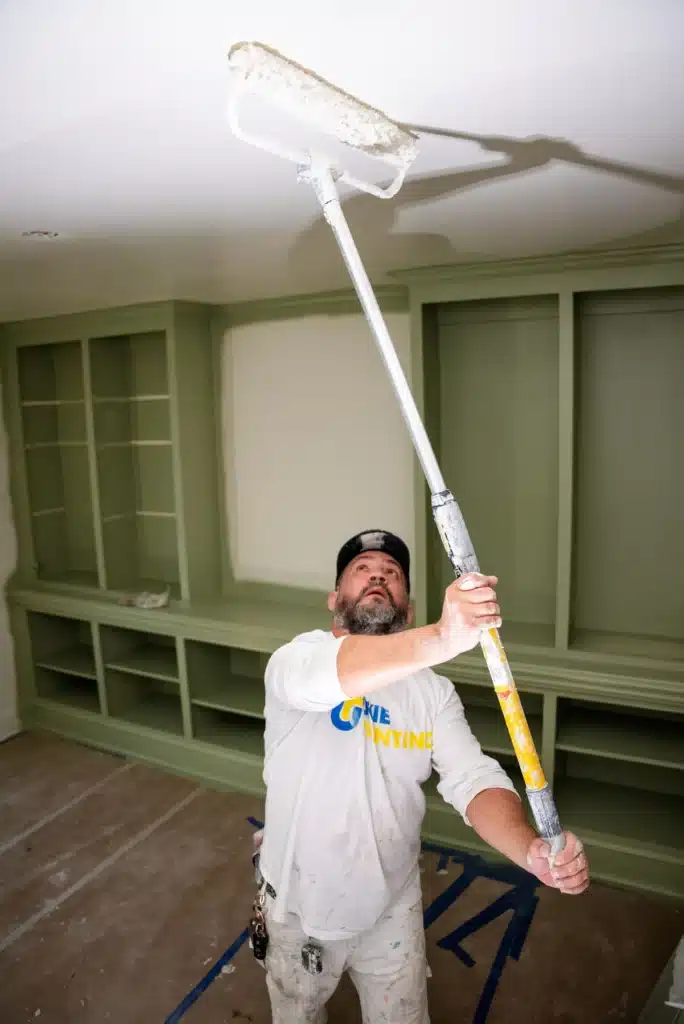 Expert Ceiling Painting for Residential Interiors A professional painter using an extension roller to paint the ceiling above green built in shelves.