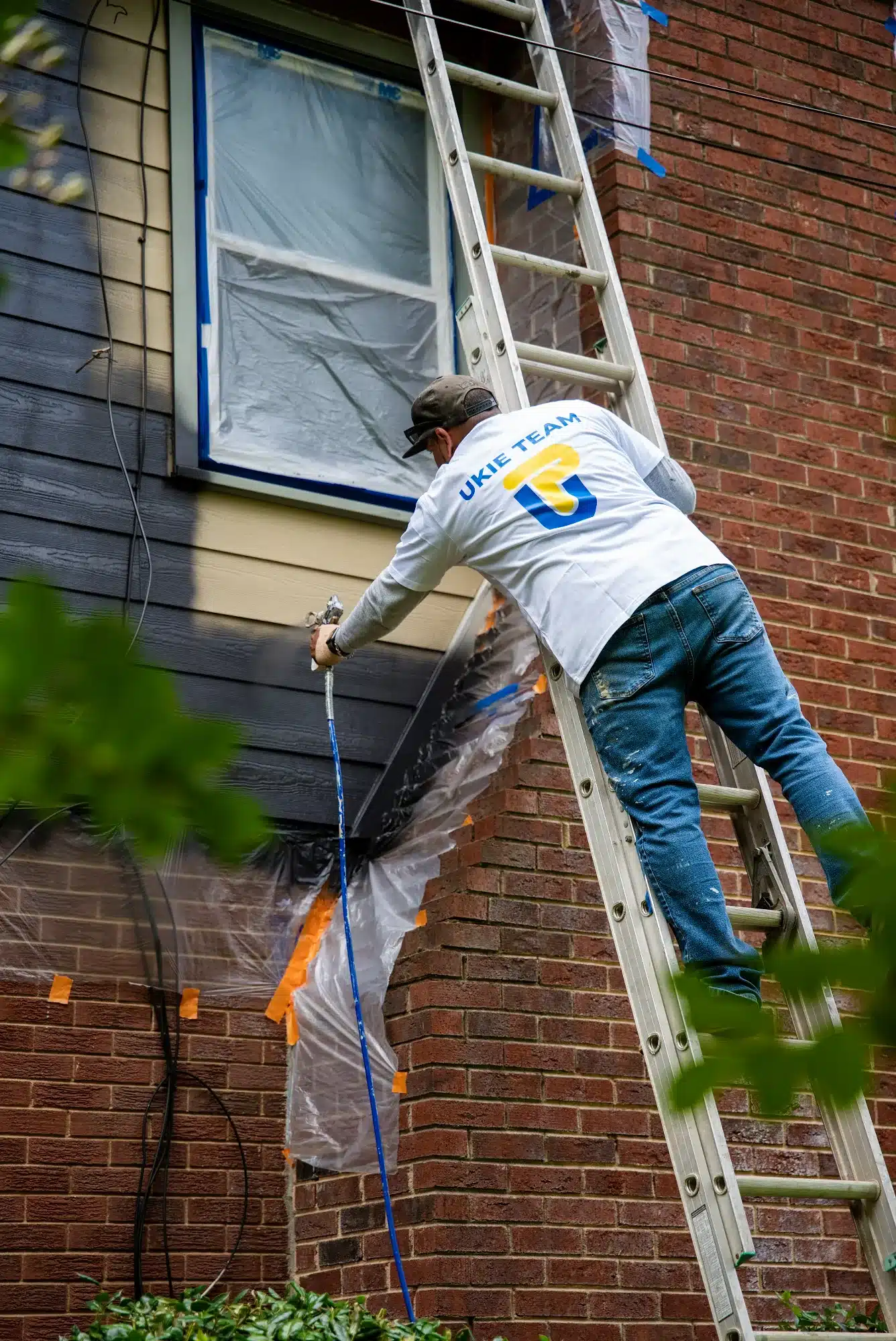 Exterior House Painting Project Painter on ladder applying paint to red brick residential exterior with blue tape and plastic sheeting protection.